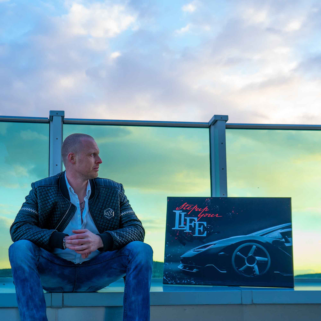 Mann sitzt neben Motivationsbild mit Sportwagen und Spruch auf einer Dachterrasse bei Sonnenuntergang