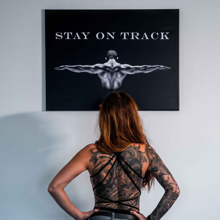Frau mit Tattoo betrachtet Motivationsbild im Gym, Leinwand mit trainiertem Mann und Schriftzug.